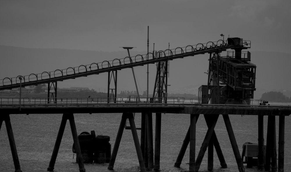 A monochrome photo of a gravel loader. From the left a jetty stands high above the water on crossed stilts. On the jetty we can see the back of a crane like object with a conveyor belt feeding into the top