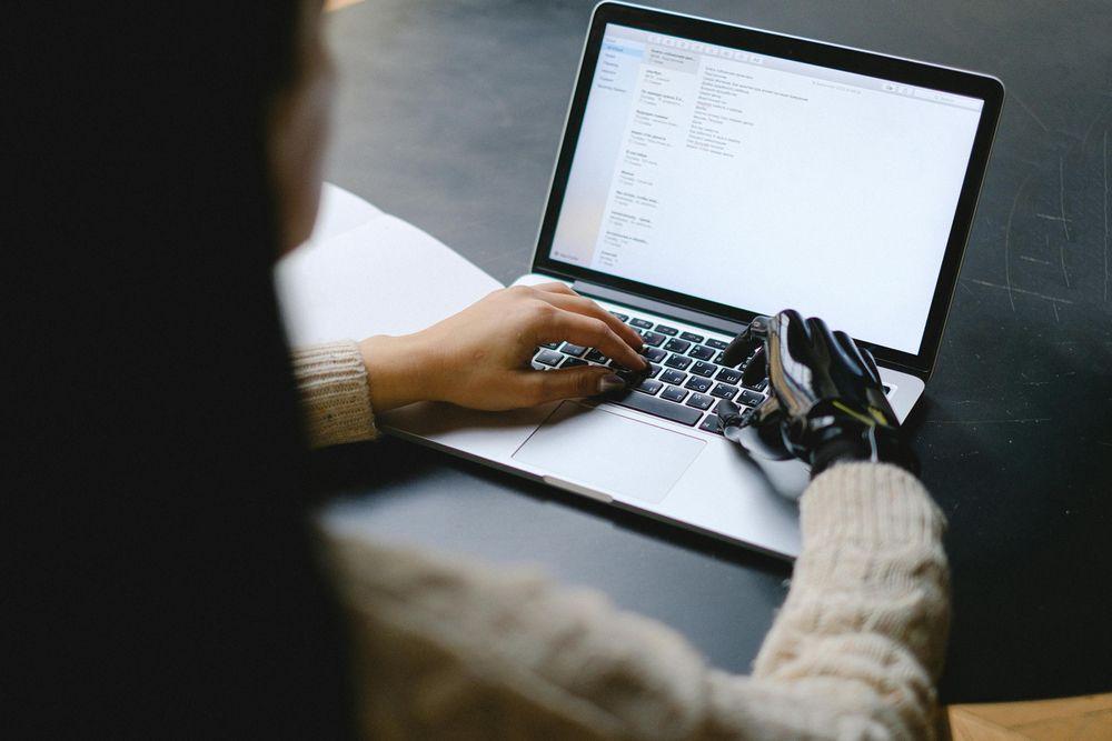 Looking over a persons shoulder towards their laptop. One hand on the keyboard is a flesh and blood hand, the other is a chrome and black prosthetic