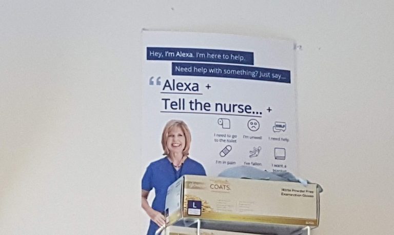 A sign on a hospital wall. It features a smiling nurse with the text &quot;Hey, I'm Alexa. I'm here to help. Need help with something? Just say 'Alexa tell the nurse...&quot; and then a range of options
