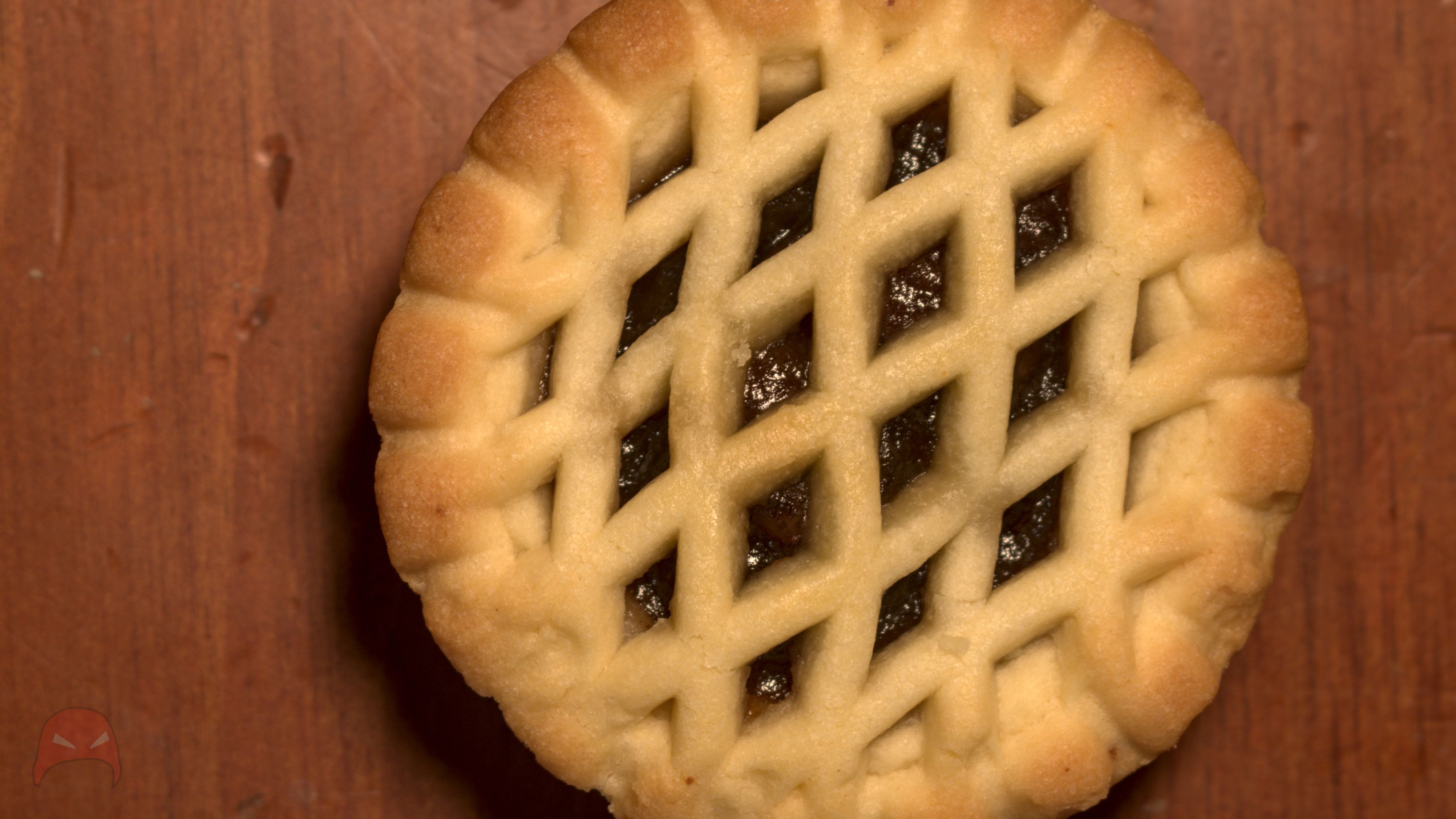A single fruit mince pies lies on a brown wooden surface. The top of the pie is covered by a lattice work of shortbread, through which you can see the fruit mince itself