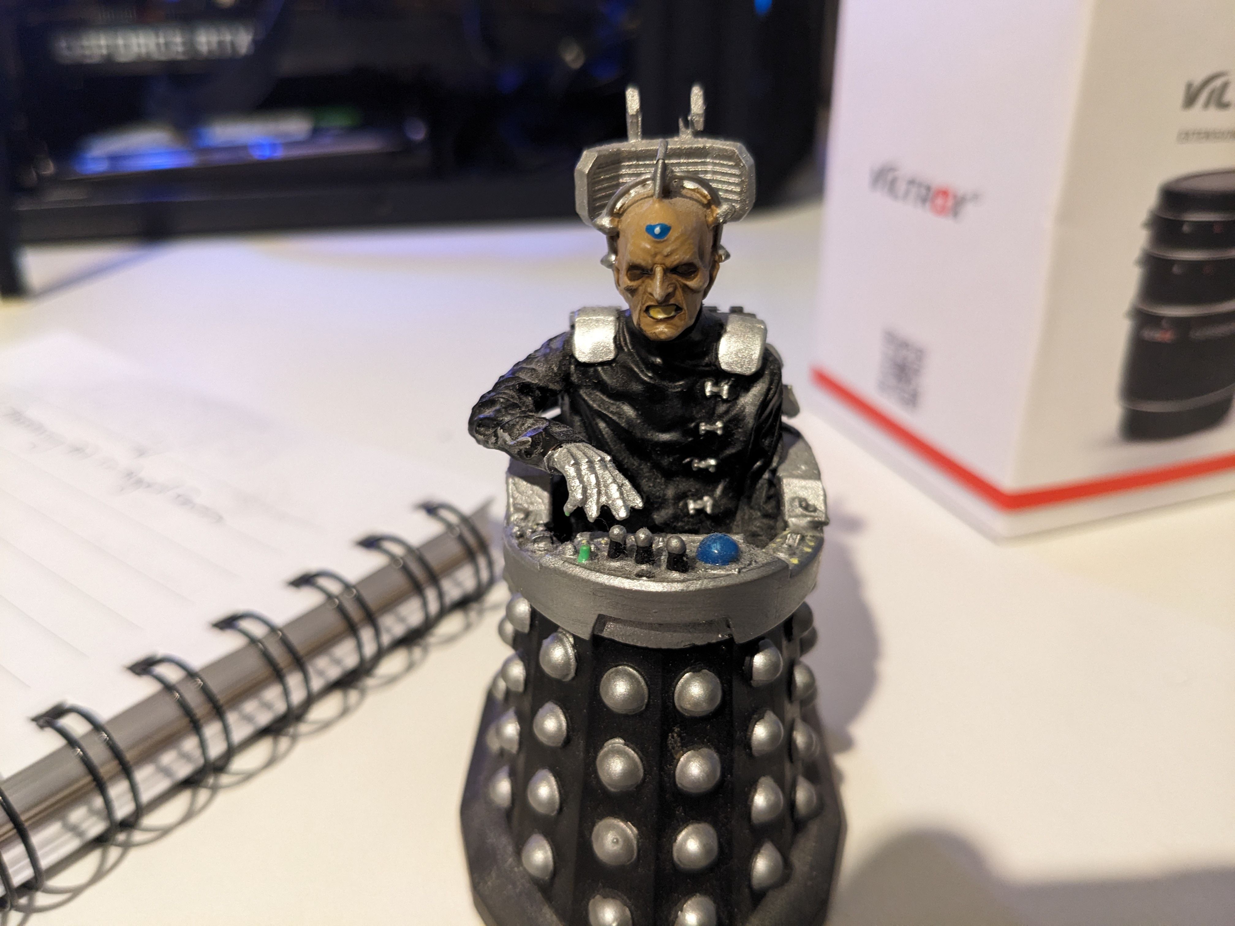 A figurine resembling a cyborg in a black and silver costume with circular patterns, in front of a notebook and a box on a desk.