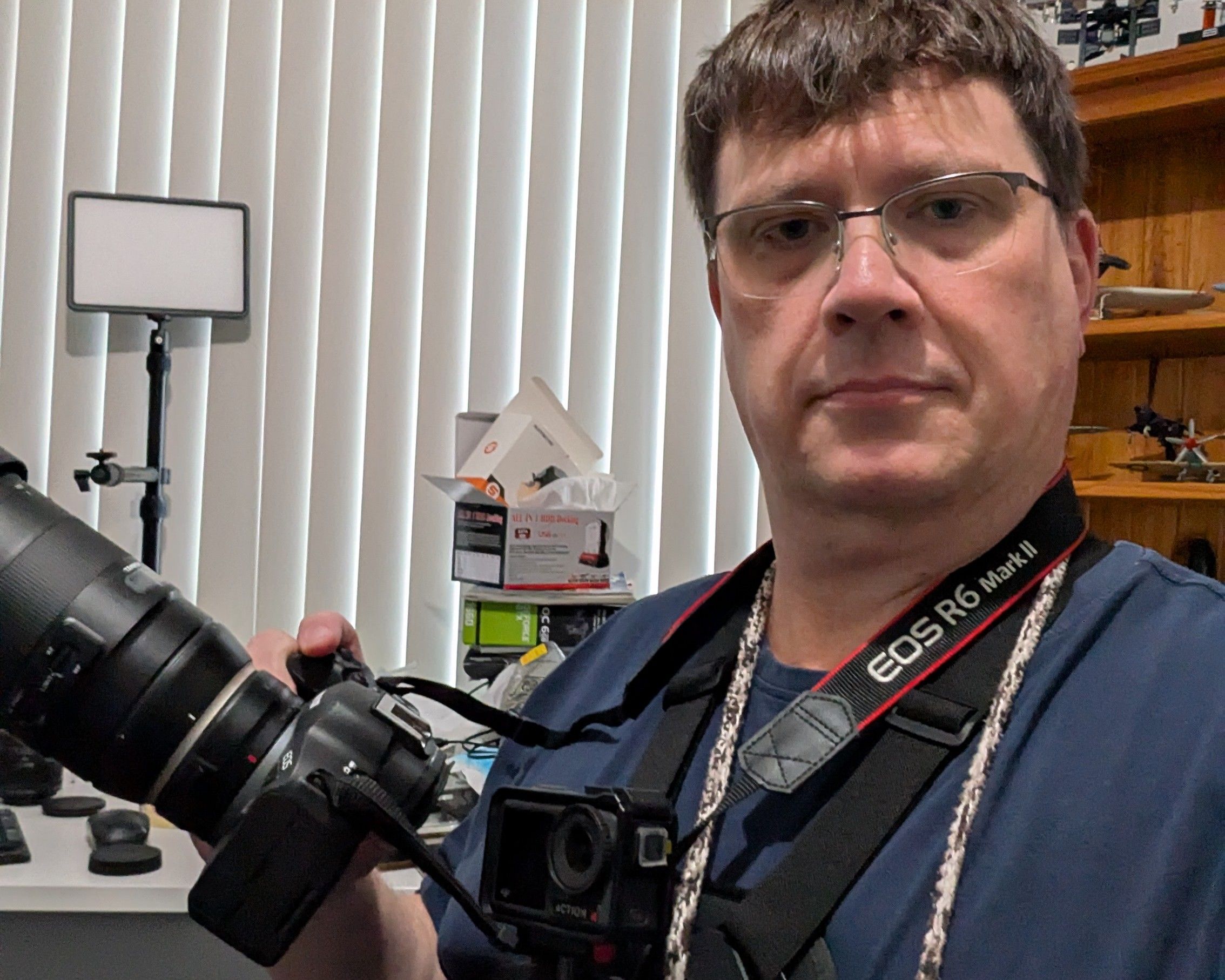 A middle aged man sits looking directly into the camera. He is holding another camera with a large lens